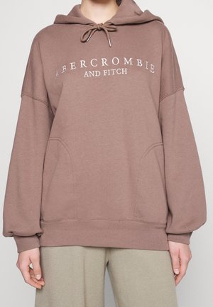 Brun oversized hættetrøje med forlommer og "Abercrombie and Fitch" tekst, båret med lyse bukser.