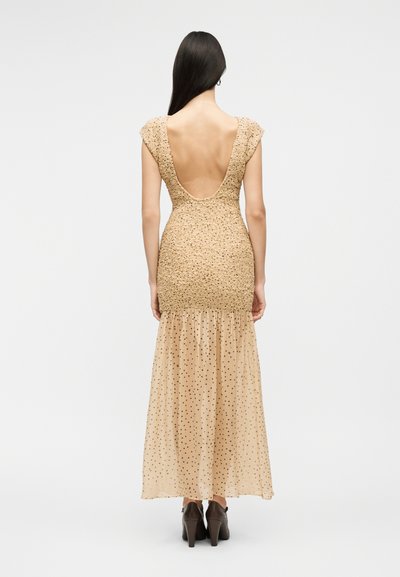 Γυναίκα με μακριά σκούρα μαλλιά, ντυμένη με μπεζ φόρεμα με ανοιχτή πλάτη, τοπ με υφή και διαφανές φούστα με πουά, στέκεται μπροστά σε απλό φόντο.