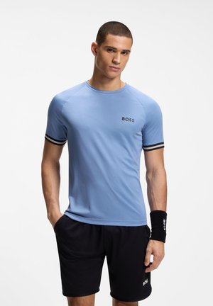 Νεαρός με κοντά μαλλιά, φορώντας ένα γαλάζιο T-shirt BOSS, μαύρα σορτς BOSS και μια μαύρη βραχιολή, στέκεται με το ένα χέρι στην τσέπη.