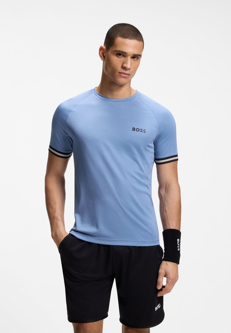 Νεαρός με κοντά μαλλιά, φορώντας ένα γαλάζιο T-shirt BOSS, μαύρα σορτς BOSS και μια μαύρη βραχιολή, στέκεται με το ένα χέρι στην τσέπη.