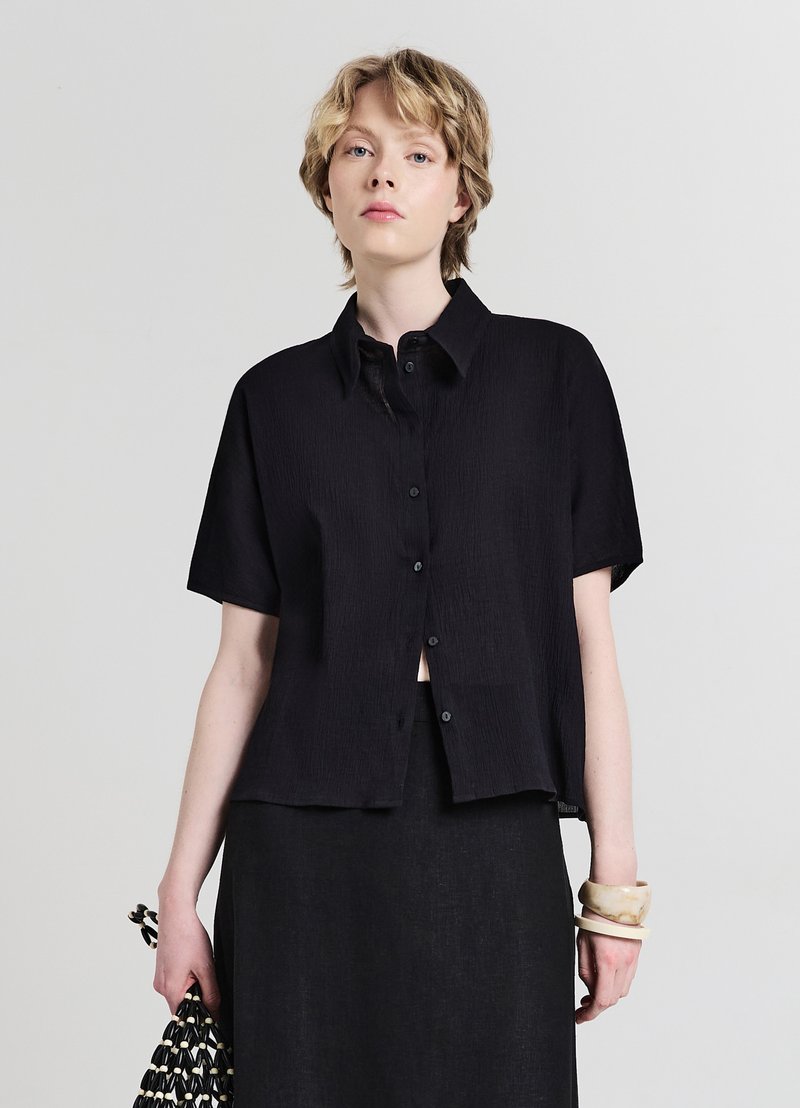 Chemise noire à manches courtes avec des boutons, en tissu texturé, col classique et coupe ample. Portée avec une jupe noire et un accessoire au poignet.