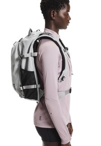 Ruksak svjetlo sive boje s crnim mrežastim umetcima, podesivim trakama i više pretinaca, koji nosi osoba u ružičastoj majici s dugim rukavima.