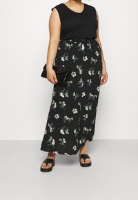 Gonna lunga nera con motivi floreali verdi e rosa, abbinata a un top nero senza maniche e una pochette nera. Presenta sandali neri flat.