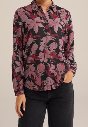 Kobieta ubrana w czarną bluzkę z długim rękawem z dużymi różowymi i bordowymi kwiatowymi wzorami, w połączeniu z ciemnymi dżinsami, dłoń w kieszeni.