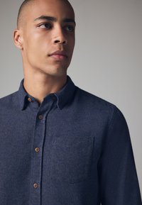 Camisa de botones azul marino con tejido texturizado, que cuenta con un cuello, bolsillo en el pecho y botones marrones. Diseño sencillo y forma clásica.