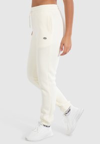 Pantalones de chándal blancos de algodón con cintura ajustable, bolsillos laterales y un pequeño parche de logo, combinados con zapatillas blancas texturizadas.