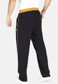 Schwarze Hose mit einem elastischen orangefarbenen Bund, schlanker Schnitt, seitlichen Taschen und orangen Paspeln entlang der äußeren Nähte.