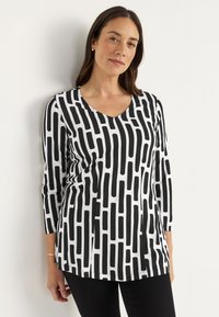 Vrouw met bruin haar, gekleed in een zwart-wit gestreepte lange mouwen top en zwarte broek, staand tegen een witte muur.
