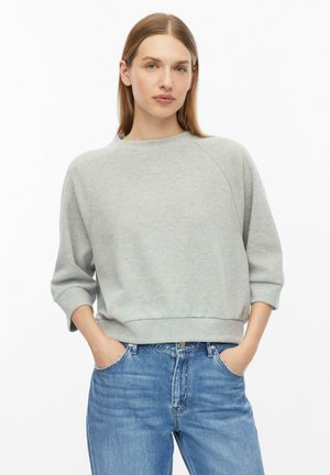 Frau mit glattem hellbraunem Haar trägt ein hellgraues Langarm-Sweatshirt und blaue Jeans mit den Händen in den Taschen vor weißem Hintergrund.