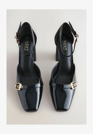 Crne lakirane visoke potpetice sa četvrtastim prstom, podešivim remenom za gležanj i detaljem u zlatnoj boji na kopči. Glatka tekstura, potpetica srednje visine.