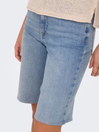 Svetlomodré rifľové šortky nosené osobou, ktorá stojí, v kombinácii s béžovým pleteným topom a zlatým prsteňom na jednej ruke.