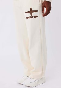 Crèmekleurige sweatpants met een gestructureerde afwerking, voorzien van een bruin geborduurd logo op het linkerbeen. Draagt met witte sneakers.