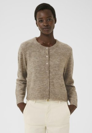 Donna indossa un cardigan beige con bottoni e pantaloni avorio, in piedi con le mani nelle tasche su uno sfondo bianco semplice.