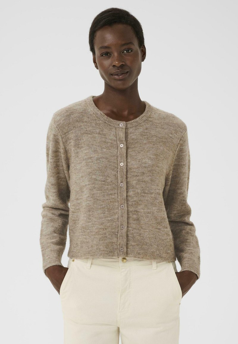 Femme portant un cardigan beige boutonné et un pantalon écru, debout avec les mains dans les poches devant un fond blanc uniforme.