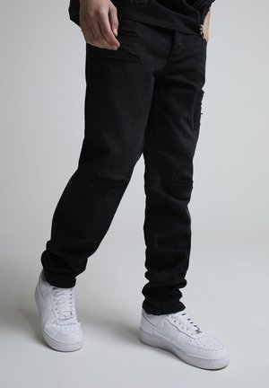 Ben iført sorte revnede jeans og hvide Nike-sneakers, stående på en lys grå gulv mod en ensfarvet baggrund.