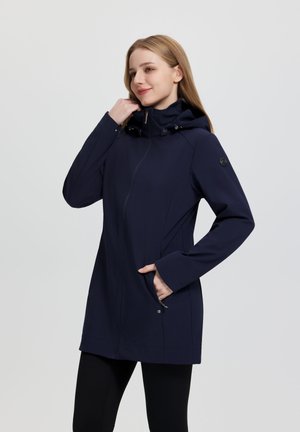 Femme portant une veste à capuche bleu marine avec une fermeture éclair à l'avant, la main dans la poche zippée, et un pantalon noir, debout devant un fond gris uni.