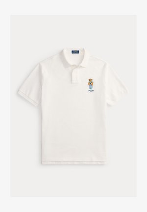 Polo bianca a maniche corte con colletto, abbottonatura a due bottoni e ricamo di un orso che indossa una giacca blu e un maglione arancione sopra la scritta "POLO".