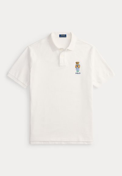 Hvit polo-skjorte med korte ermer, krage, to-knapps plastrong og brodert bjørn som har på seg blå jakke og oransje genser over teksten "POLO".