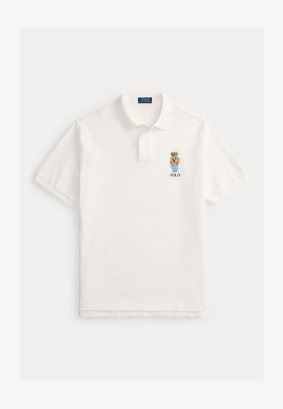 Hvit polo-skjorte med korte ermer, krage, to-knapps plastrong og brodert bjørn som har på seg blå jakke og oransje genser over teksten "POLO".