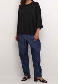 Schwarzes, locker sitzendes Oberteil mit weiten Ärmeln, kombiniert mit blauen Jeans. Die Schuhe sind schwarze Blockabsatzsandalen.