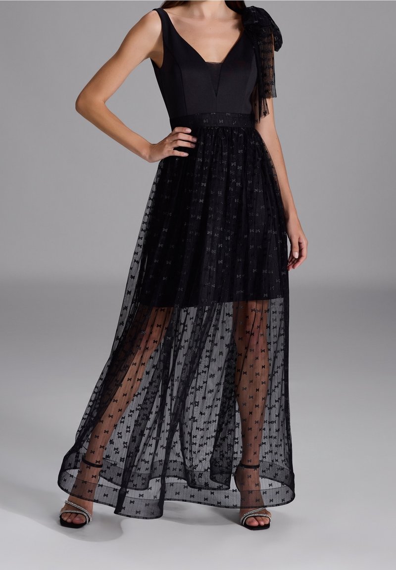 Femme portant une robe noire sans manches avec un voile transparent à motifs et un nœud sur une épaule, associée à des talons ouverts noirs.