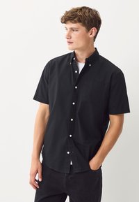 Camicia nera a maniche corte con bottoni, realizzata in cotone, dotata di colletto, tasca singola sul petto e bottoni bianchi a contrasto. Indossata sopra una t-shirt bianca.