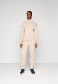 Veste à zip beige et joggers assortis en tissu doux. La veste présente un col montant et une poche kangourou ; les joggers ont une coupe décontractée.