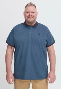 Sötétkék pólóing sötét gallérral, két gombos légkörrel és rövid ujjakkal. A mellen kis logó található. Khaki nadrággal viselve.