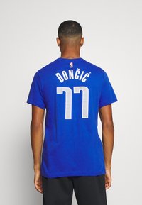 Uomo di spalle che indossa una maglia NBA blu con "DONČIĆ" e il numero 77 in bianco sulla parte posteriore, su uno sfondo semplice.