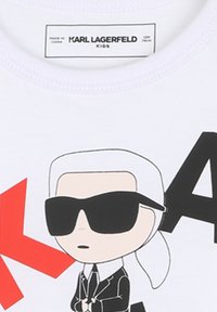 T-shirt bianco in cotone con un personaggio dei cartoni animati che indossa occhiali da sole, un vestito nero e capelli bianchi, arricchito da elementi grafici rossi. Etichetta del marchio ben visibile.
