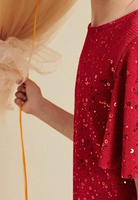 Robe rouge à manches courtes recouverte de paillettes brillantes. Une main tient un tissu léger et une corde orange. Apparence texturée et festive.