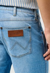 Jeans de mezclilla celeste con un parche de cuero marrón de Wrangler en el bolsillo trasero, con costuras y remaches amarillos que contrastan.
