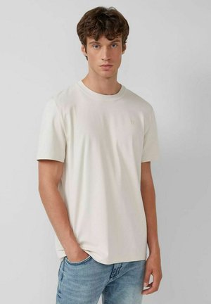 Junger Mann mit lockigem braunem Haar, der ein einfaches weißes T-Shirt mit kurzen Ärmeln und hellblauen Jeans trägt, steht vor einem grauen Hintergrund.