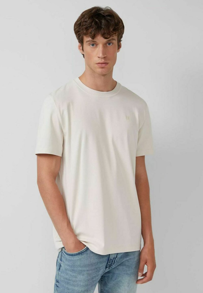 Young man with curly brown hair wearing a plain white short-sleeve t-shirt and light blue jeans, standing against a gray background.