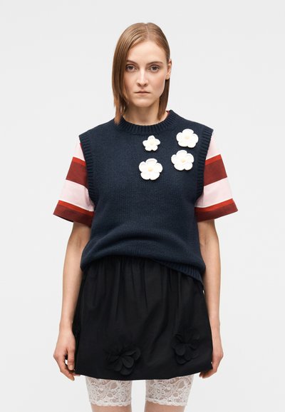 Pull en tricot bleu marine avec applications de fleurs blanches, associé à une chemise à manches courtes rayée et une jupe noire ornée d'accents floraux.