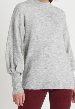 Vrouw draagt een lichtgrijze gebreide trui met pofmouwen en geribbelde manchetknopen, gecombineerd met donkerrode broek.