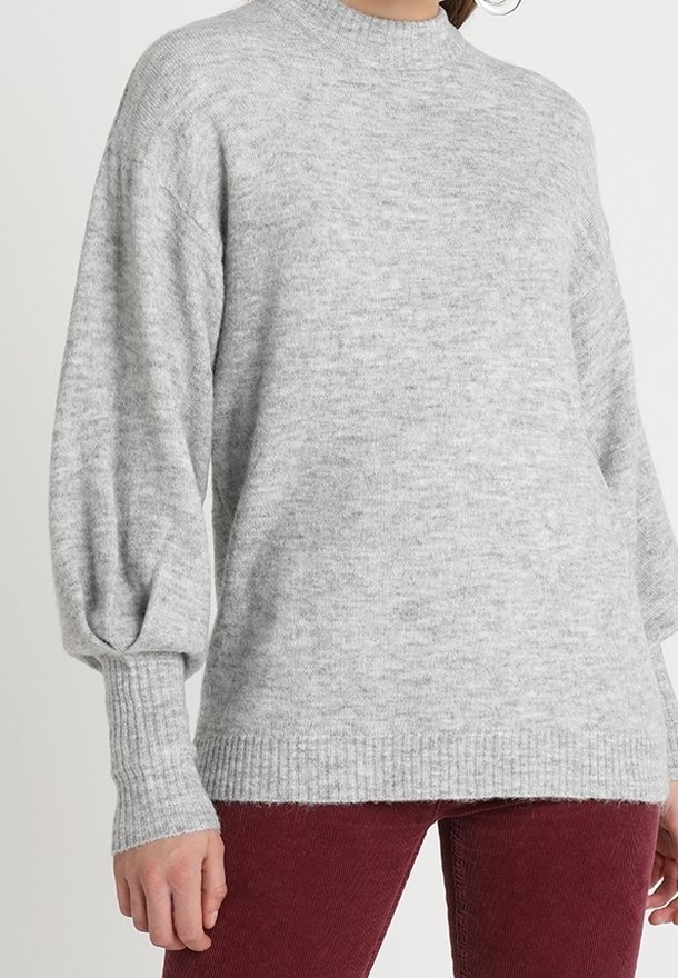 Femme portant un pull en maille gris clair avec des manches longues bouffantes et des poignets côtelés, assorti à un pantalon rouge foncé.