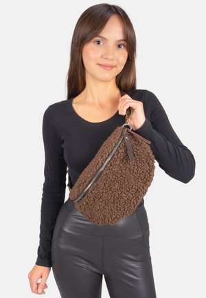 Braune Texturtasche mit einem weichen, flauschigen Außenteil, ausgestattet mit einem silbernen Reißverschluss und einem eleganten schwarzen Tragegurt, gehalten von einer Person in einem schwarzen Oberteil.