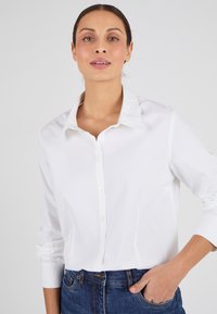 Camisa blanca de botón con cuello, de mangas largas y una textura suave y ligera. Combinada con jeans de mezclilla oscuros, mostrando un estilo clásico.