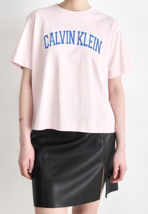 Osoba nosící světle růžové tričko Calvin Klein s modrým nápisem a černou koženou sukni s předním rozparkem.