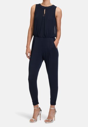 Combinaison en bleu marine avec un design sans manches, décolleté en forme de trou de serrure, taille élastique, poches latérales et jambes fuselées. Le modèle porte des sandales à talons hauts argentées.