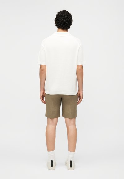 Άνδρας με σγουρά μαλλιά, που φοράει λευκό πουκάμισο με κοντά μανίκια, ελιά πράσινες βερμούδες, λευκές κάλτσες και λευκά αθλητικά παπούτσια, στέκεται γυρισμένος προς τα πίσω.