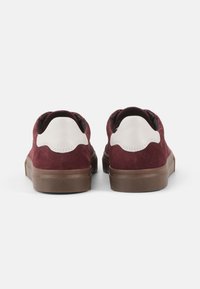 Burgundy suede sneakers with a white leather accent at the heel, featuring a smooth brown rubber sole and rounded shape.