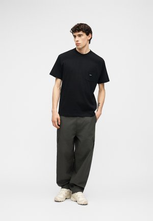 Νεαρός άνδρας που στέκεται με τα χέρια στις τσέπες, φορά μαύρο μπλουζάκι με τσέπες, σκούρο γκρι φαρδιά παντελόνια και λευκά αθλητικά παπούτσια σε απλό φόντο.