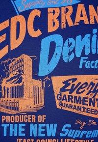 T-shirt in cotone di colore blu navy con stampa grafica arancione e blu, che presenta un'illustrazione di un edificio e un testo sulla produzione e garanzia del denim.