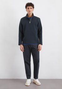 Mörk marinblå kvartdragpullover i texturerad tyg, matchad med mörka byxor och krämfärgade sneakers. Har en stående krage och sidofickor.