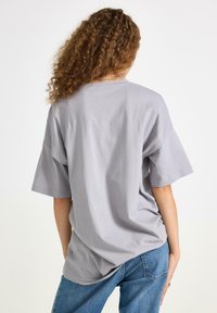 Jasnoszary, oversize t-shirt z krótkim rękawem i okrągłym dekoltem. Wykonany z miękkiego, bawełnianego materiału, w luźnym kroju, włożony w niebieskie dżinsy.
