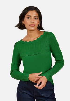 Kvinde iført en grøn langærmet sweater med flosset halsudskæring og mørkeblå bukser, stående mod en ensfarvet baggrund.