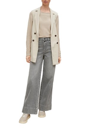 Blazer sans manches beige sur un haut beige clair, associé à un jean gris large et des baskets beiges, présentant des accents de boutons et une texture subtile.