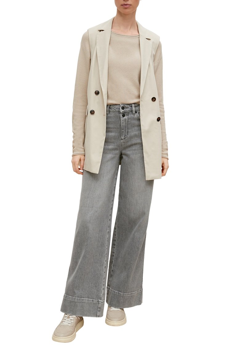 Beige ärmelloser Blazer über einem hellbeigen Oberteil, kombiniert mit weiten grauen Jeans und beigen Sneakers, die über Knopfdetails und eine subtile Textur verfügen.